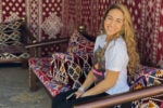A person with long hair smiling and sitting on a decorated couch with colorful geometric patterns and a similarly patterned backdrop.