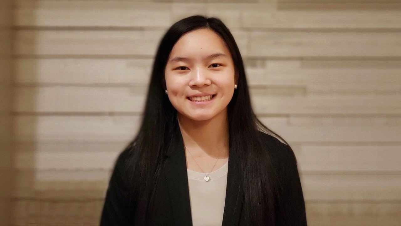 A headshot of a smiling person with long dark hair wearing a black blazer and a necklace, standing against a beige background.