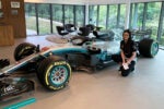 A person kneeling next to a Formula 1 race car in a modern showroom with large windows and lounge furniture. The car features logos and branding.