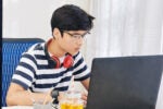 A person wearing glasses and headphones is focused on a laptop, seated at a desk with a container of yellow liquid and a straw in front.