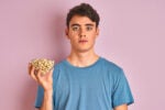A person holding a bowl of pistachios in one hand, wearing a blue shirt, set against a plain pink background.