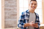 A person smiling and holding a smartphone, wearing a checkered shirt over a gray t-shirt, standing indoors near large windows.