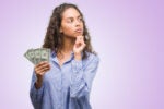 A person with curly hair holding dollar bills while looking thoughtfully to the side, set against a purple background, suggesting financial contemplation.