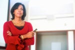 A person in a red blouse holding a presentation clicker, standing in a bright room, possibly a lecture or conference setting.