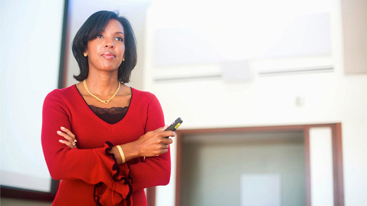 A person in a red blouse holding a presentation clicker, standing in a bright room, possibly a lecture or conference setting.