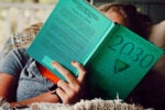 A person lies on a couch, reading a teal-colored book titled "2030." The reader''s fingernails are painted orange, and they are wearing a smartwatch. The environment appears cozy and relaxed.
