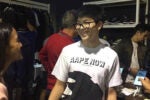 A group of people inside a clothing or shoe store. One person in the foreground is wearing glasses and a T-shirt with "AAPE NOW" printed on it, smiling and talking to another person. Shelves of shoes are visible in the background.