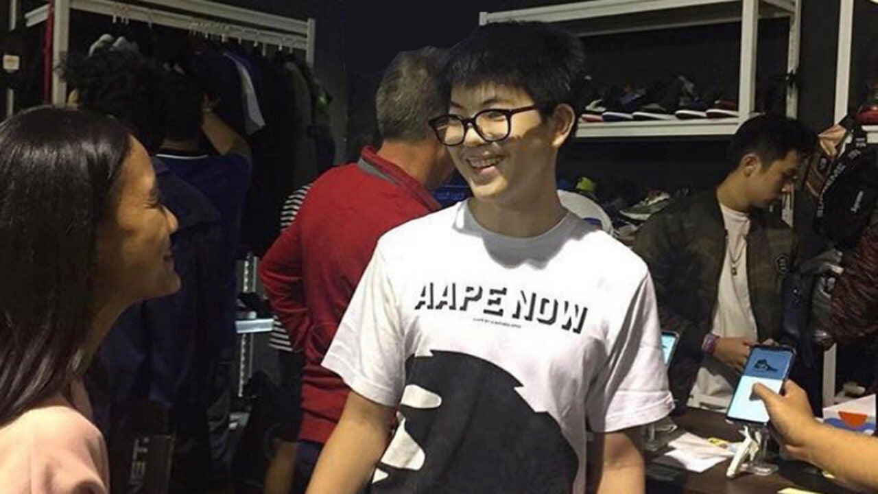 A group of people inside a clothing or shoe store. One person in the foreground is wearing glasses and a T-shirt with "AAPE NOW" printed on it, smiling and talking to another person. Shelves of shoes are visible in the background.