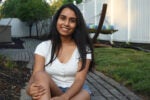 A person with long hair is smiling and sitting on a stone pathway in a backyard. There''s a hammock and a white fence in the background.