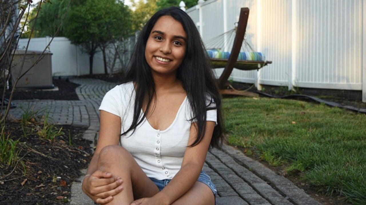 A person with long hair is smiling and sitting on a stone pathway in a backyard. There''s a hammock and a white fence in the background.