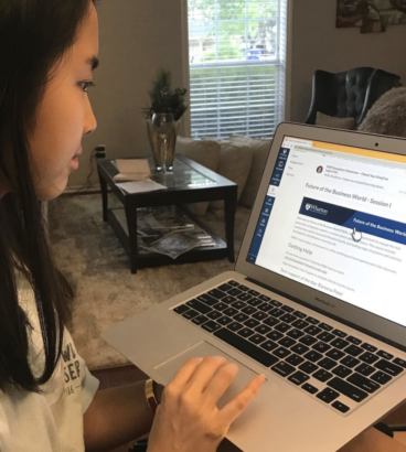 A person using a laptop in a living room setting, viewing online educational content titled "Future of the Business World - Session 1" from Wharton, with a window and coffee table in the background.