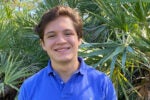 A person smiling, wearing a blue polo shirt, stands in front of lush green palm leaves.