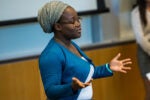 A person in a blue jacket and headscarf is speaking or presenting in a classroom or conference setting, with gestures indicating engagement.