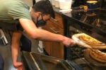 A person wearing a mask and apron is using a wooden pizza peel to place a pizza into an oven.