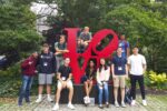 A group of people posing around a large red "LOVE" sculpture in a garden setting.