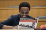 A person smiling while using a laptop in a classroom. The laptop is adorned with stickers, including one that says "I eat data for breakfast."