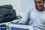 A person is smiling while using a laptop, next to a backpack and a travel mug on a table.