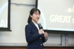 A person in a suit is speaking into a microphone in front of a presentation screen displaying partial text "GREAT".