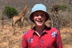 A smiling person wearing a blue bucket hat and a red floral shirt stands in the foreground, while a giraffe is visible in the background on a dry, grassy landscape.