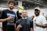 Three people posing in a casual setting, with one holding up a peace sign and another a rock hand sign. A person in the background displays a bright shoe. The environment suggests an indoor event.