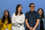 Four people are standing together in front of a blue background, smiling and appearing cheerful.