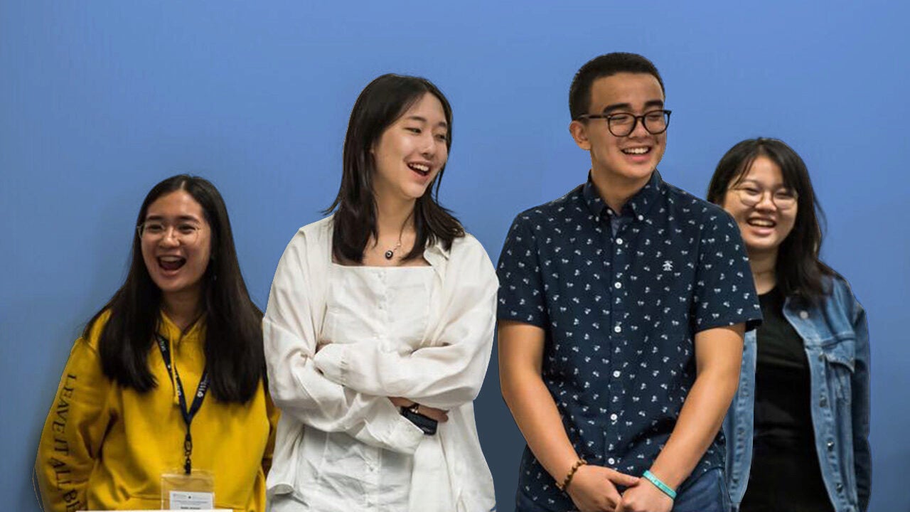 Four people are standing together in front of a blue background, smiling and appearing cheerful.