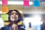 Person thoughtfully looking at colorful sticky notes on glass, suggesting brainstorming or planning in an office setting.