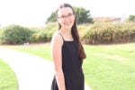A person with long hair and glasses, wearing a black sleeveless dress, smiles while standing on a path in a sunlit park with greenery.