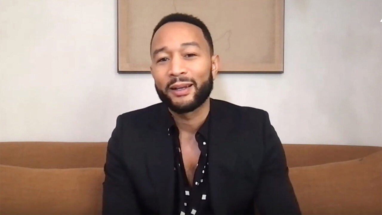 A person with short hair and a beard, wearing a black jacket and patterned shirt, sitting on a couch against a plain background.