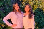Two people in light-colored sweaters stand together in front of lush green bushes, smiling at the camera with sunlight casting warm tones.