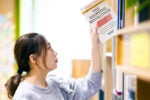 A person reaching for a book titled "The Shopping Revolution" on a bookshelf. The setting suggests a library or bookstore.
