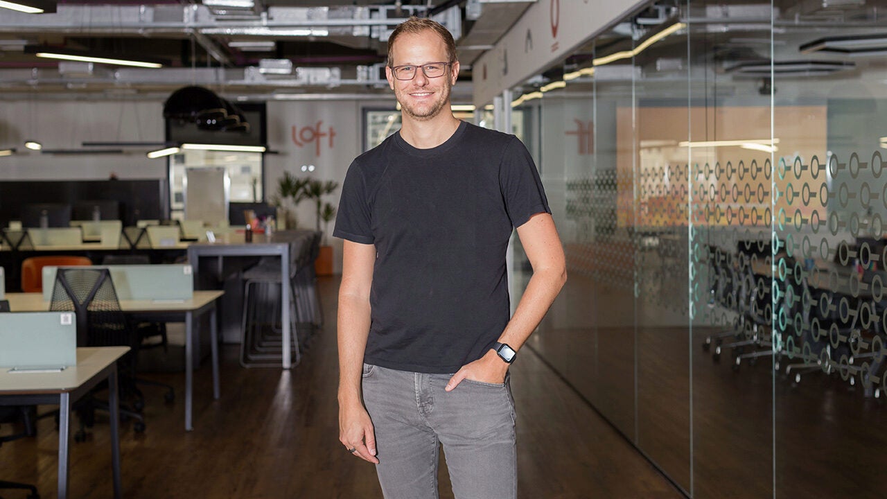 A person standing in a modern office space with glass walls and desks, wearing a black t-shirt and jeans, smiling at the camera.