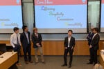 A group of people in business attire are giving a presentation in front of a projected screen displaying the words "Core Ideas: Efficiency, Simplicity, Convenience, Accessibility."