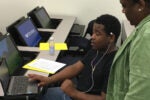 A student sitting in a classroom, engaged with a laptop and wearing earbuds, while another person stands nearby giving assistance. Desks with additional computers and papers are visible.