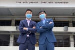 Two individuals wearing suits and masks stand with arms crossed. A sign above them reads "Live with passion Lead with compassion."