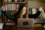 A person with long curly hair is sitting at a desk with a laptop, raising their arms in excitement. Bookshelves are in the background.