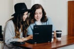 Two people sitting at a wooden table, looking at a laptop. One wears a black hat. There are two coffee cups beside them.