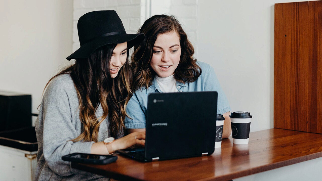 Two people sitting at a wooden table, looking at a laptop. One wears a black hat. There are two coffee cups beside them.