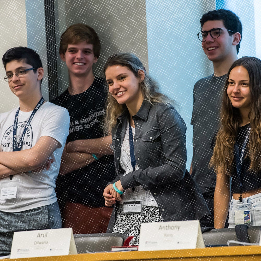 A group of five smiling individuals stands casually against a wall, wearing name tags, suggesting they are at a conference or workshop.