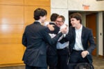 A group of four people in suits celebrating and hugging in a room with wooden paneling and an exit sign.