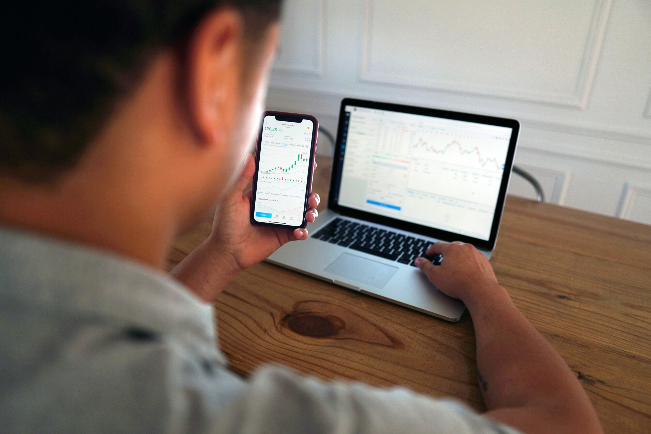 A person analyzing stock market charts on a smartphone and laptop at a wooden table.