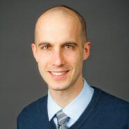 Headshot of a person with short hair wearing a blue sweater, light blue shirt, and striped tie, with a neutral background.