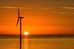 Silhouette of an offshore wind turbine at sunset over calm water, with the sun near the horizon and a vibrant orange sky.