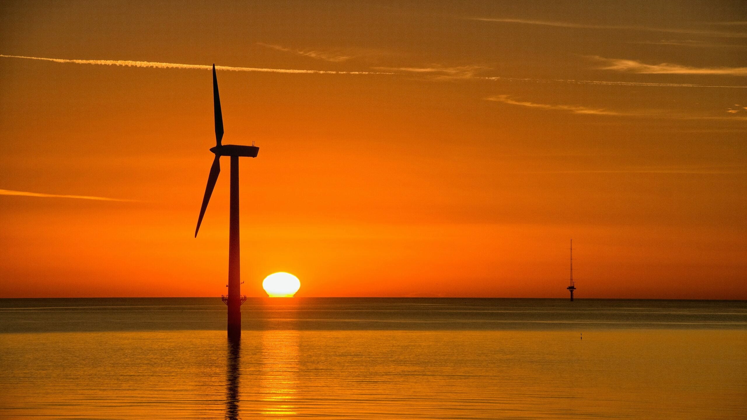 Silhouette of an offshore wind turbine at sunset over calm water, with the sun near the horizon and a vibrant orange sky.