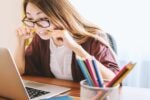 A person with glasses biting a pencil while looking at a laptop, conveying stress or concentration, with colored pencils in a cup nearby.