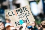A protest sign reading "Climate Justice Now!" with a thermometer and Earth illustration, held by a person in a crowd.