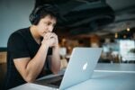 A person wearing headphones is concentrating on a laptop in a lightly blurred indoor setting, suggesting focus or online work.