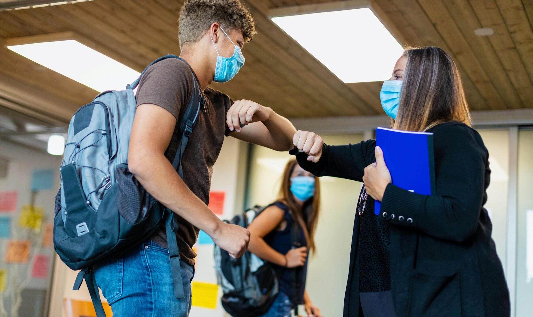 A student and a teacher wearing masks exchange an elbow bump in a classroom. The student has a backpack, and the teacher holds a folder.