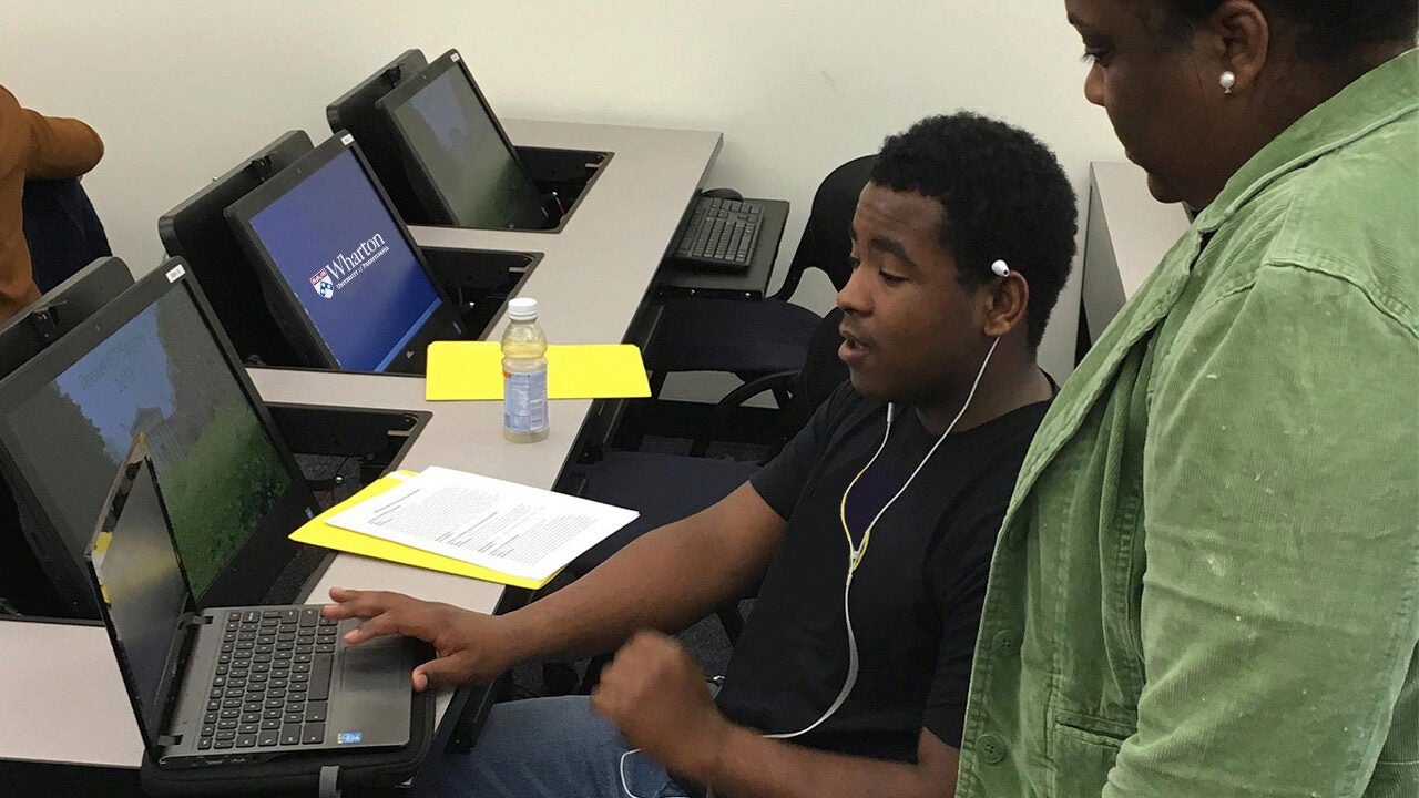 A person seated at a desk using a laptop in a classroom, engaged in conversation with another person standing nearby. Laptops are visible on adjacent desks.