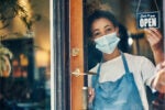 A person wearing a face mask and apron stands at a door, holding an "Open" sign, indicating a business is ready for customers.
